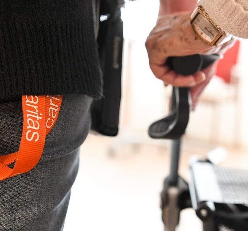 Ältere Frau hält sich am Rollator fest, während Caritas-Fachkraft sie begleitet.